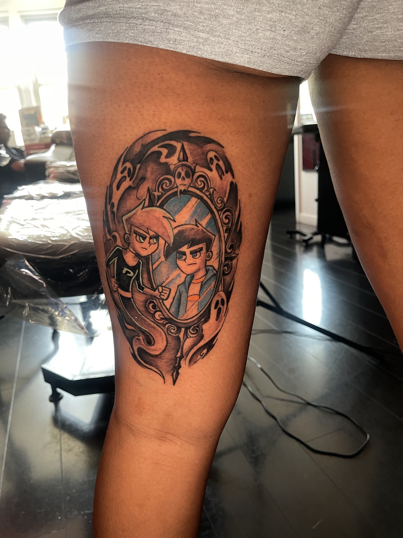 New school color tattoo of two cartoon characters framed in an ornate haunted mirror, on a client's thigh